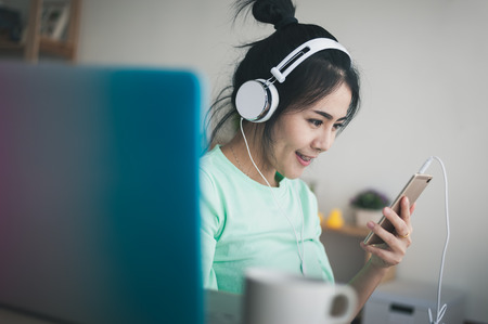 Young attractive Asian woman listening music on smartphone while working with laptop computer. Internet of things for modern lifestyle and freelance worker conceptの写真素材