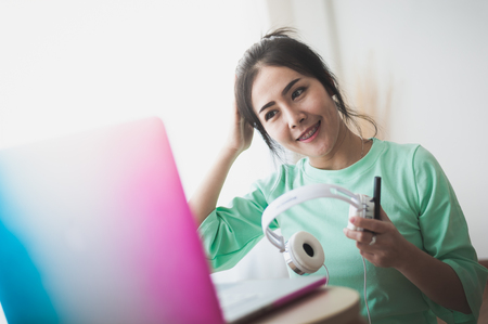 Young attractive Asian woman taking headphone off while working with laptop computer. Internet of things for modern lifestyle and freelance worker conceptの写真素材