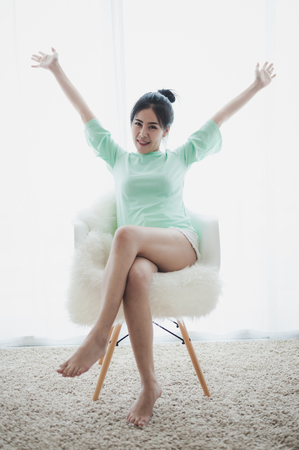 Young attractive Asian woman wear green casual cloths raise her arms up while sitting on chair in livingroom on weekendの写真素材