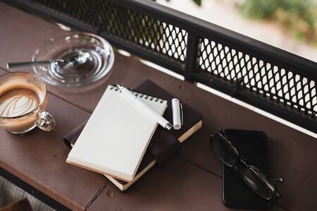 Abstract scene of opened small notebook, pen, glasses, mobile phone, and coffee cup on wood counter.の写真素材