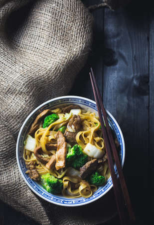 A big bowl of asian beef noodles with broccoli and cabbage in a dark setting with chopsticksの写真素材