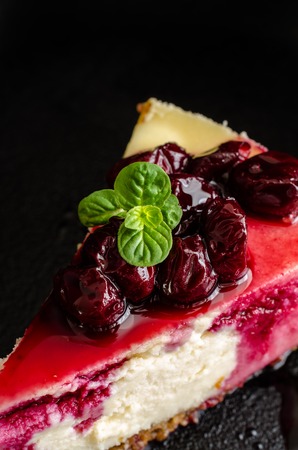 A piece of cheesecake decorated with cherry sauce with berries and a mint leaf on black plateの写真素材