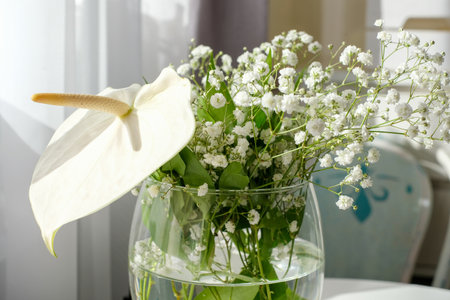 A beautiful, large, white anthurium flower and fragile, delicate flowers in a white bouquet in a glass vase. Sun rays in white curtains in the background. copyspace. high quality photoの写真素材