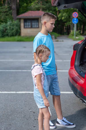 A girl with braids is standing interestedly next to the boy by the trunk of a red car with a parking in the background. High quality photoの写真素材