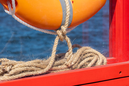 A lifebuoy is attached to a bright orange wooden frame with linen ropes, its section visible. Dark...