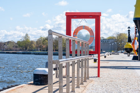Metal tube construction, lifebuoy in red wooden frame between the sea and the pavement. Metal equipment, building and green trees in background. Copy space. Background for quotes. High quality photoの写真素材