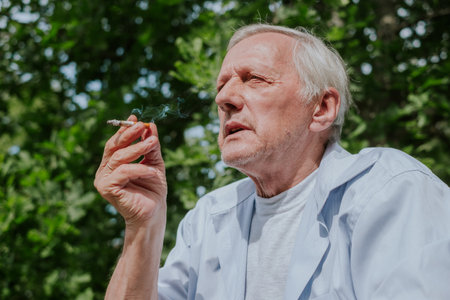 Old man smoking a cigarette in a sunny summer day, candid shot suitable for discussions about lifestyle and aging, unhealthy habits and addictions, insurance ad. High quality photoの写真素材