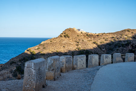 A sunlit path curves towards a whitewashed building on a headland overlooking the deep blue sea, guarded by weathered bollards. High quality photoの写真素材