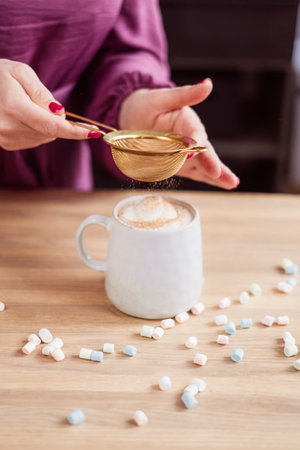 Hands delicately dusting cocoa over a frothy cup of cappuccino, with scattered marshmallows on the table, embodying a moment of cafe like indulgence at home. High quality photoの写真素材