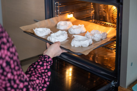 A person in a patterned shirt slides a tray of meringues into the oven, an intimate moment in home baking, perfect for illustrating recipe steps or baking classes. High quality photoの写真素材