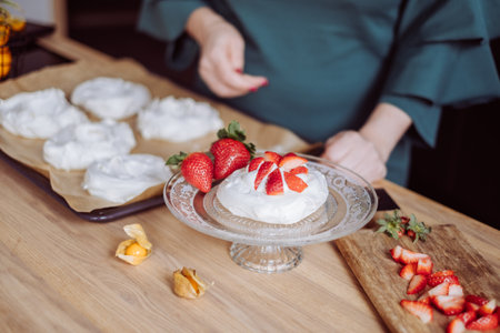 A chef in a teal apron presents a beautifully assembled strawberry pavlova on a glass stand, with more meringues and freshly sliced strawberries laid out on a wooden table. High quality photoの写真素材