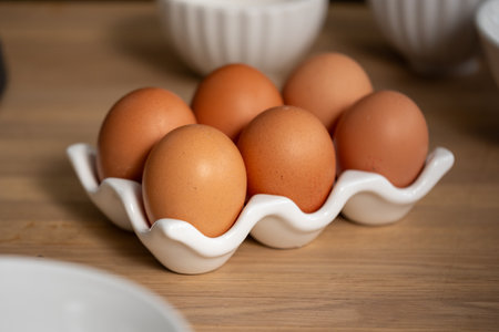 A close up view of a white ceramic egg holder on a wooden table, neatly cradling a row of fresh brown eggs, set amidst other baking ingredient, everyday cooking elements. High quality photoの写真素材