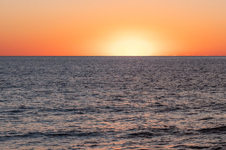 The sun sinks below the horizon, creating a stunning display of orange and yellow tones. The tranquil ocean reflects the fading light, enhancing the evening tranquility.の写真素材