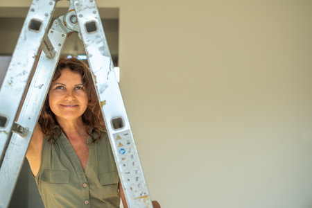 Confident midlife woman stands next to a ladder, smiling in a well-lit room with neutral walls,...