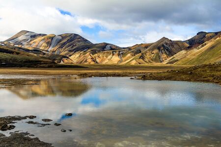The rainbow rock of Landmannalaugar is reflected in a still lake under a cloudy sky, Iceland.の写真素材