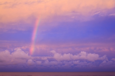 A bright rainbow arches out of a pink sea into a dusky pink and purple magenta sky, framed by magenta cloudsの写真素材