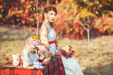 Beautiful bride with bouquet in hands sitting in the autumn park near the table with cups and flowersの写真素材