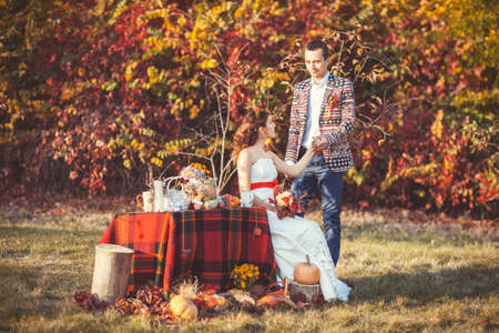 Bride sitting near the table in autumn park the groom standing side by side and holding her handの写真素材