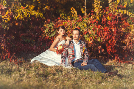 Beautiful bride in a wedding dress and groom sitting near the bushes in the autumn forestの写真素材