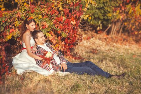 Groom is sitting on the grass in the autumn park leaning against a brideの写真素材