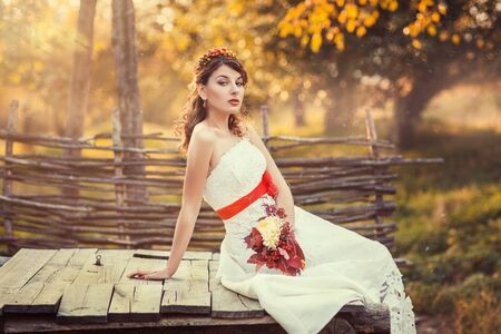 Beautiful bride sitting on the closed wooden well in the autumn parkの写真素材