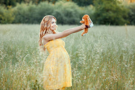 The pregnant redhaired girl standing in a field holding a toy in front of herself looking at toy and smilingの写真素材