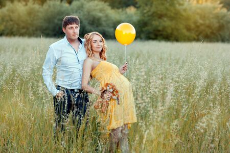 Young man and his pregnant wife standing in the field and looking into the distance. The girl is holding a toy and balloonの写真素材