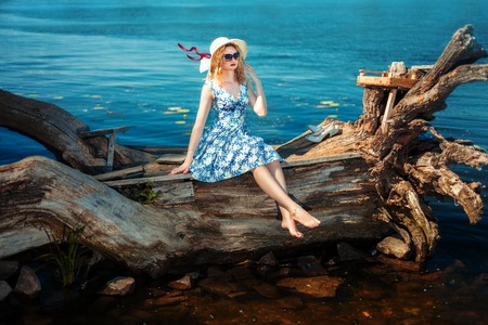 Blonde girl in a hat and sunglasses sitting on driftwood on the riverbank. She put her hand to her faceの写真素材