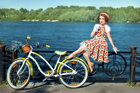 The girl is sitting on the fence on the river bank. Next to her is a bicycle. The bike basket with flowersの写真素材