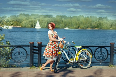 Beautiful girl standing near a bicycle on the river bank and looks away. The bike basket with flowersの写真素材