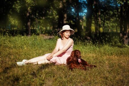 Beautiful girl in a hat sitting on the meadow and looking at lying next to her dogの写真素材