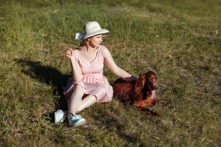 Beautiful girl in a hat sitting on the meadow. Next to her is a dog