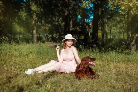 The girl in a hat sitting on the meadow. Next to her is a dog. Dog stuck out her tongueの写真素材