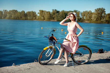 Beautiful girl in hat and sunglasses on a bicycle on the river bank. The girl looks into the distanceの写真素材