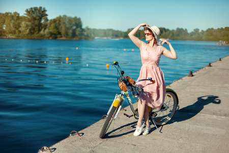 Beautiful girl in hat and sunglasses on a bicycle on the river bank. The girl looks into the distance and holds a hatの写真素材