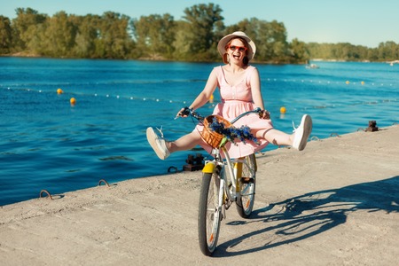 Portrait of a beautiful girl in a hat and glasses on a bicycle. She rides a bicycle on the riverbank with her legs apart. Girl laughsの写真素材