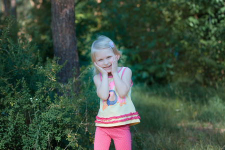 Little beautiful blonde girl standing in the forest. She girl put her face in her hands and bowed her head, she laughsの写真素材