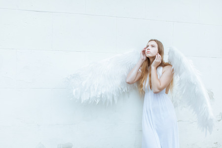 Beautiful angel girl with white wings near the wall. She looks up thoughtfullyの写真素材