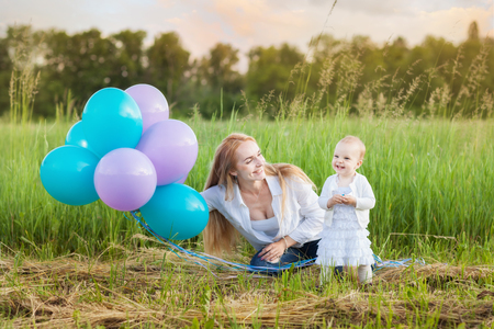 Young mother and her daughter sitting on the grass. Mum holds balloonsの写真素材