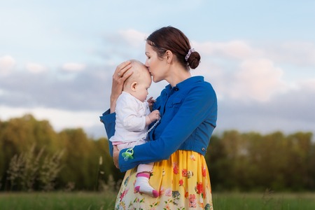 Mom holds daughter on hands. They stand in a meadow. Mother kissing daughterの写真素材