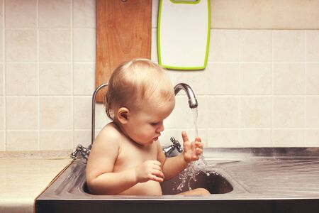 Baby is sitting in the kitchen sink and touched the water from the tapの写真素材