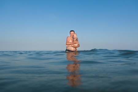 Young dad standing in the water in the sea with his daughter in his armsの写真素材