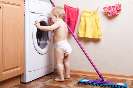 Baby girl stands near a washing machine and and looks into itの写真素材