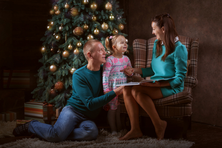 Young family near the Christmas tree. Mum reads the book to her daughterの写真素材