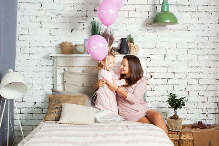 Mom and daughter are sitting on the bed. Little girl holding balloons in handsの写真素材