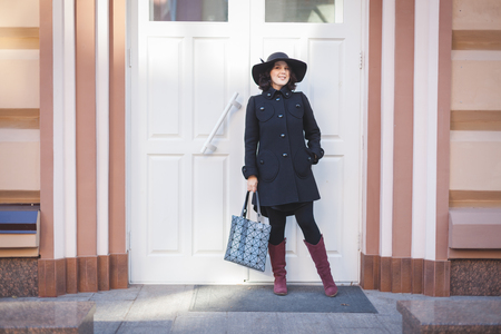 Cheerful young woman in a coat and hat stands near a white door.の写真素材