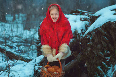 A little girl in a red coat and headscarf holds a basket of pies in her hands. She stands in the winter forest.の写真素材