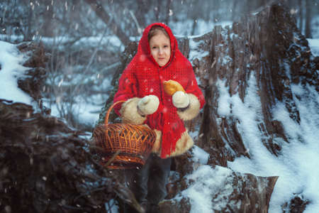 Cute girl stands in the winter forest and eats a pie. She has a basket in her hands.の写真素材