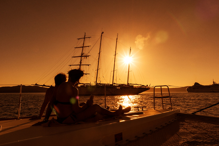 couple sitting on the bow of boat with view of the sea and sailboat, sunset, Greece, Santorini.の写真素材
