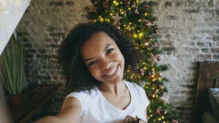 Funny mixed race girl taking selfie pictures on smartphone camera at home near Christmas treeの写真素材